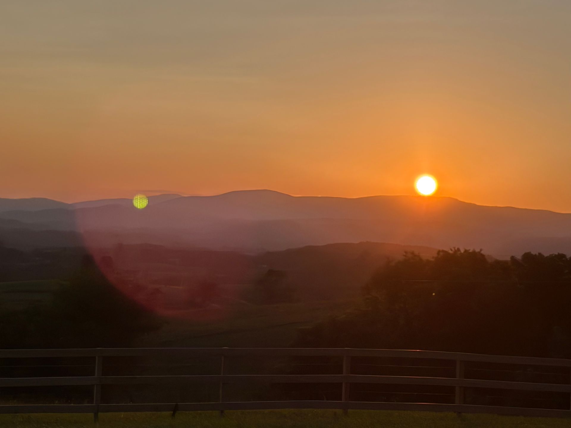 Sunset over layered mountains with silhouetted trees and a wooden fence in the foreground