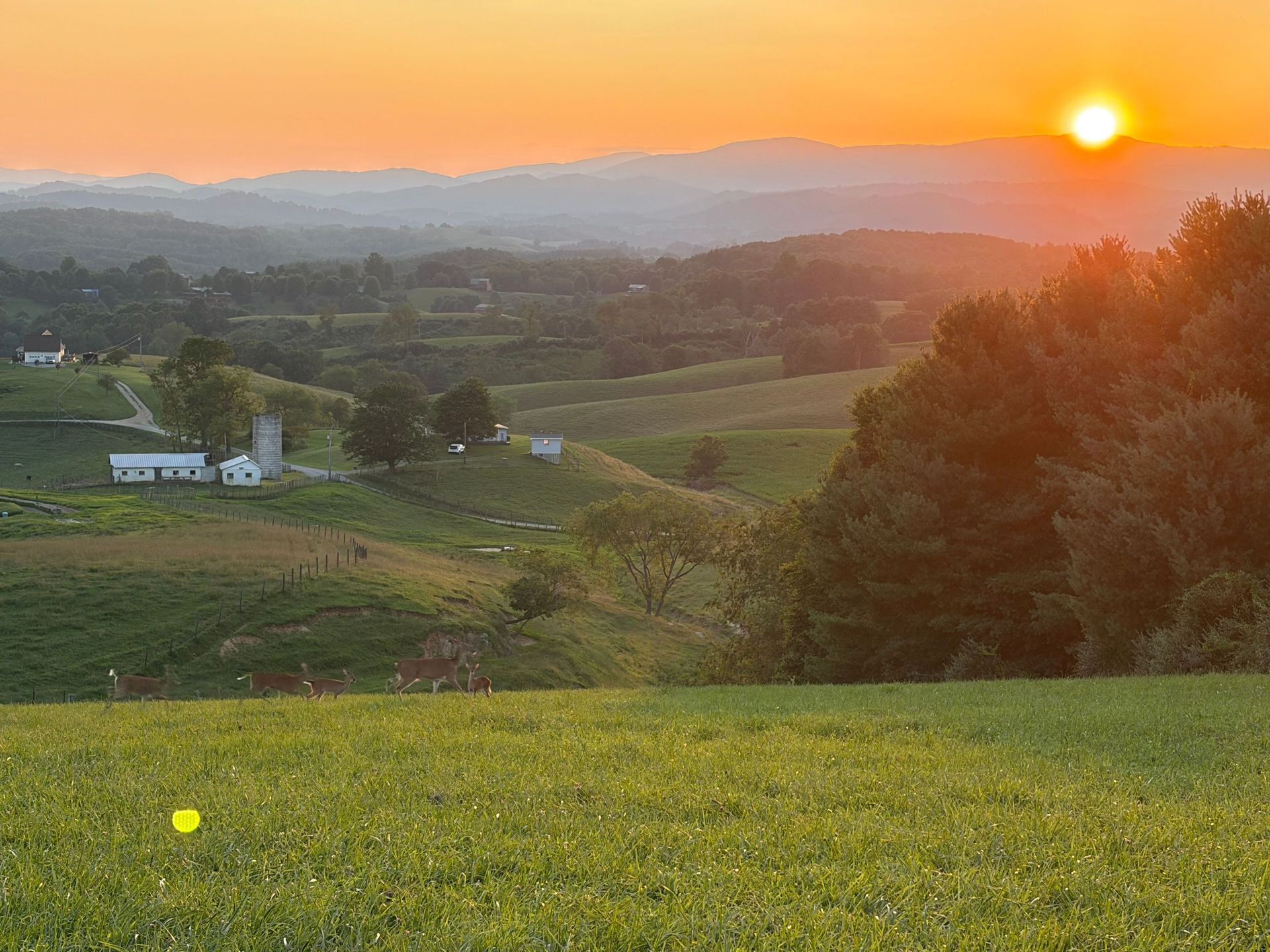 Sunset over rolling green fields and distant hills, with a farmhouse and glowing orange sky
