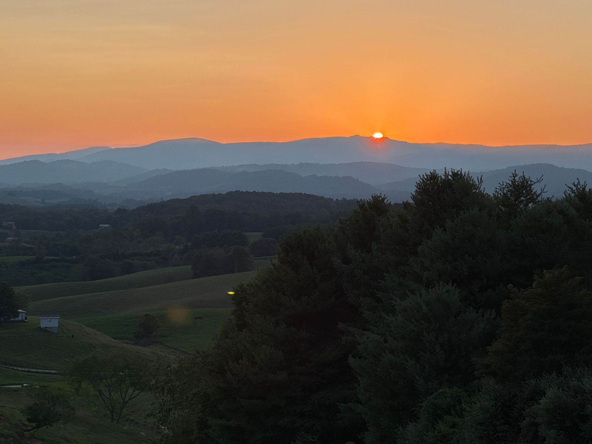 Sunset over layered hills and a dark valley, with orange sky and silhouetted trees in the foreground