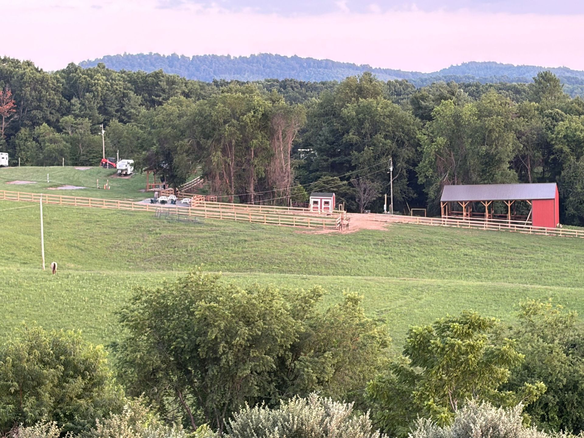 Wide green field with trees, a small red barn, and a distant wooded hillside under a pale sky