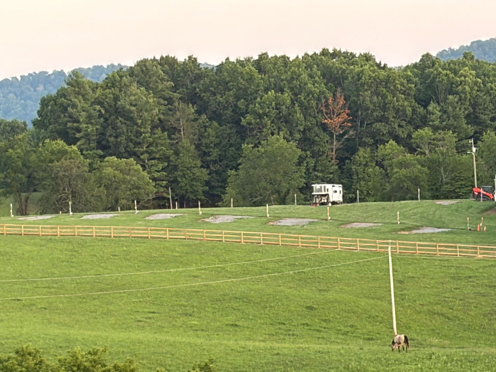Open grassy field with white fences, a small white shed, and trees along a hilltop edge