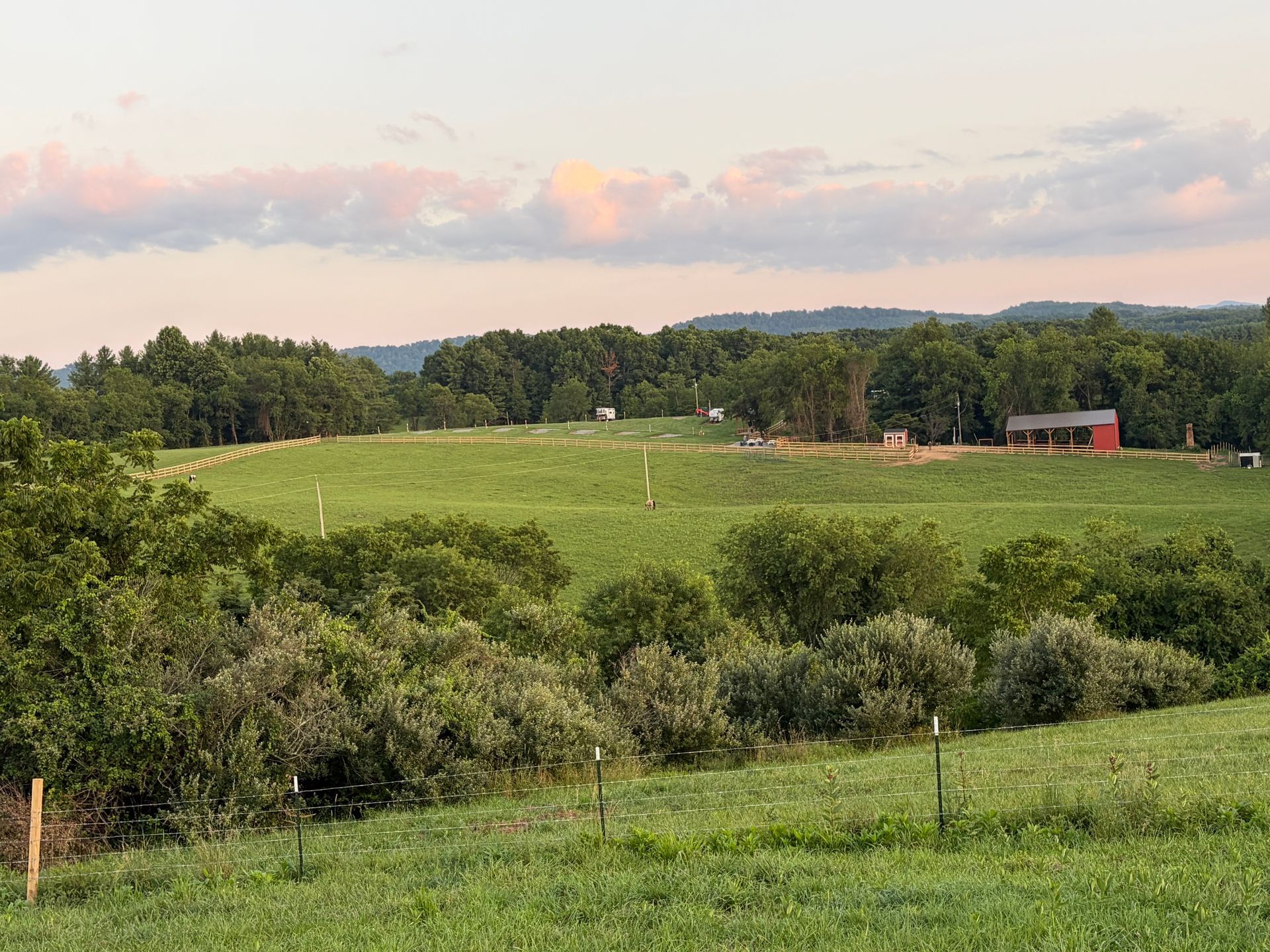 Rolling green pasture with trees, fence posts, and a red barn under a cloudy evening sky