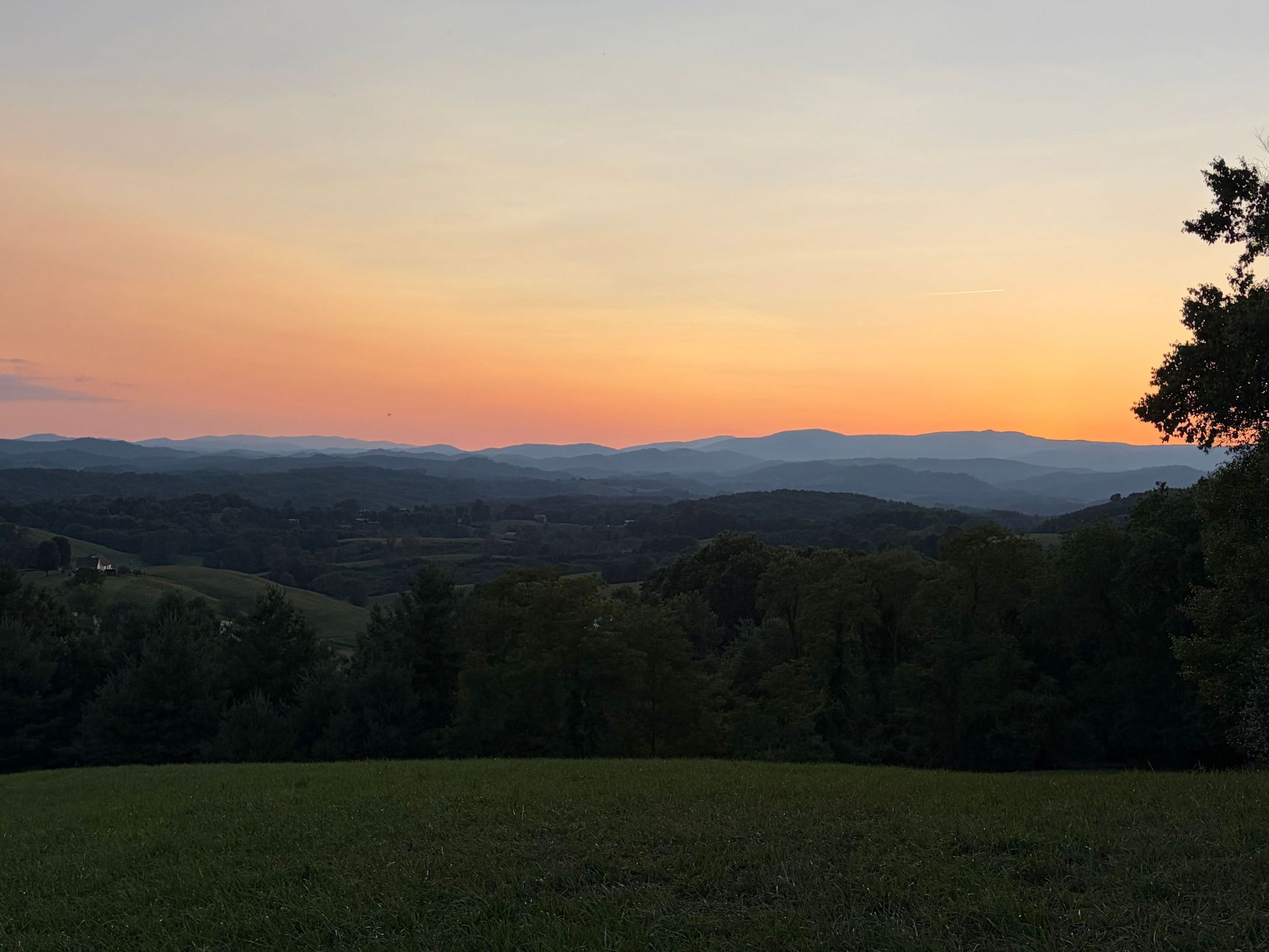 Sunset over layered mountains and a dark green meadow beneath a pink-orange sky