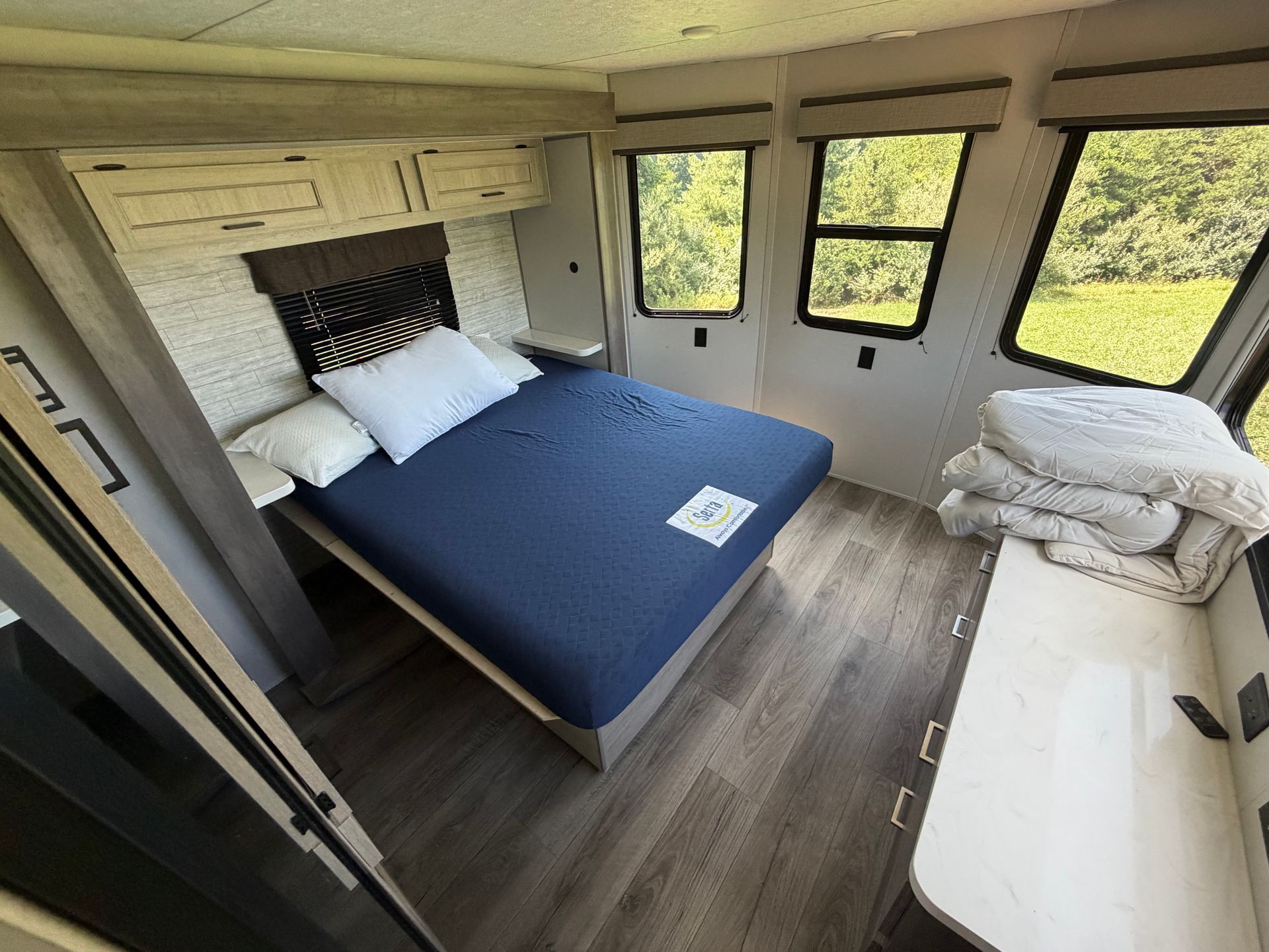 Compact camper bedroom with blue bedspread, built-in cabinets, and windows overlooking greenery