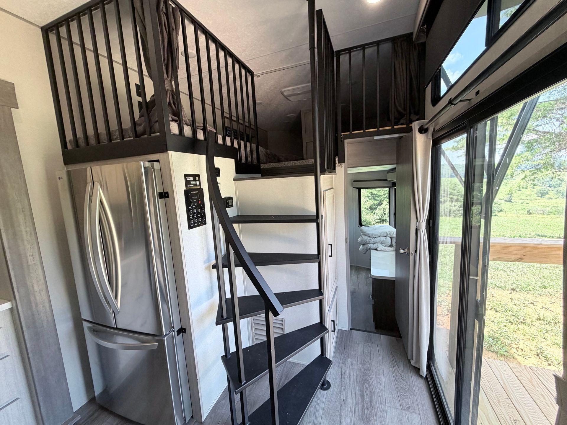 Compact RV interior with spiral stairs, stainless fridge, and large window beside a grassy view