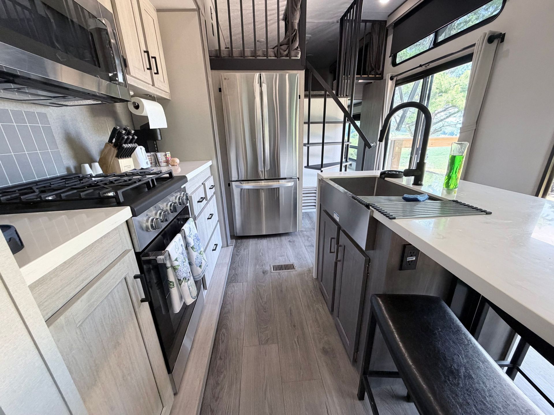 Modern galley kitchen with stainless steel appliances, white counters, and a black bench by a window