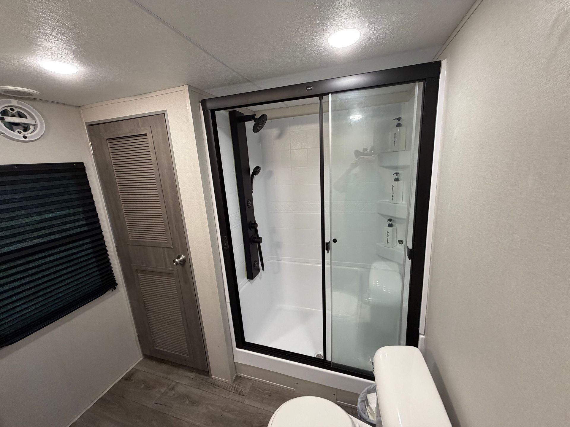 Small bathroom with walk-in shower, toilet, wood door, and dark window blinds.