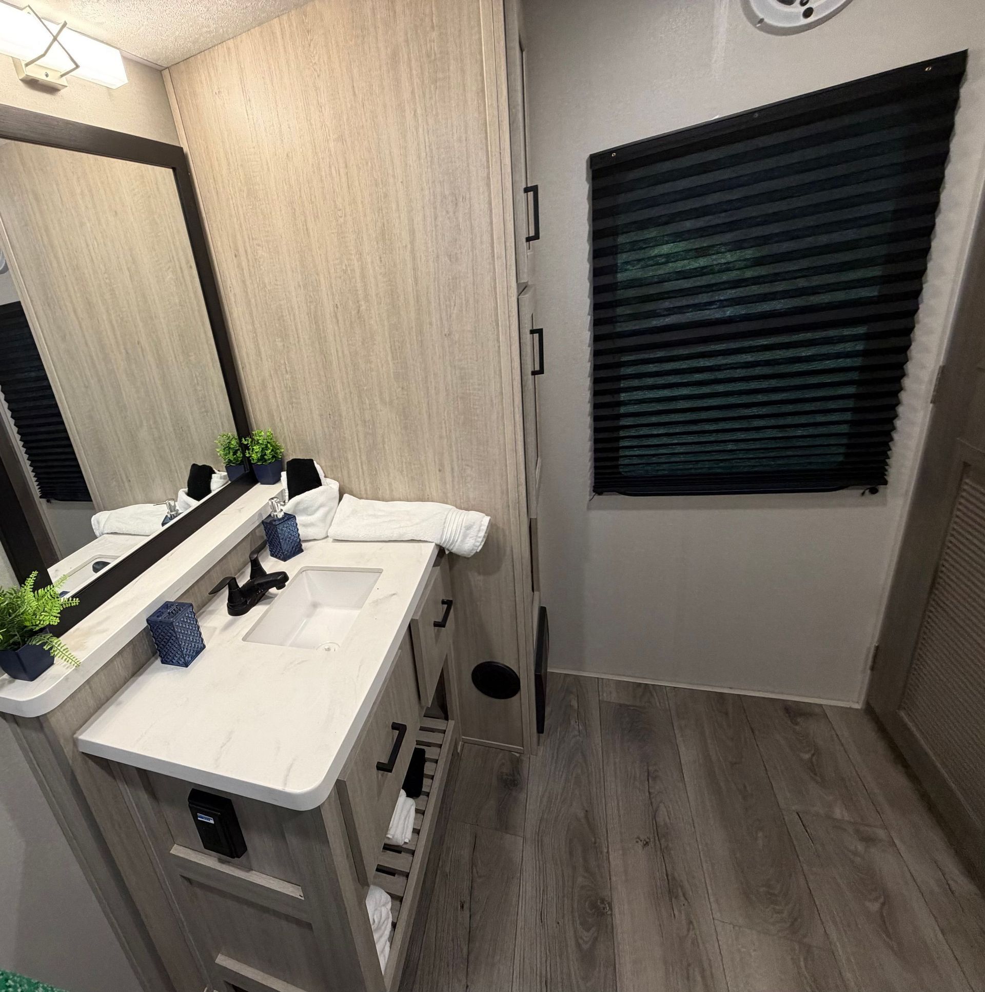 Small bathroom vanity with sink, mirror, black blinds, and light wood flooring.