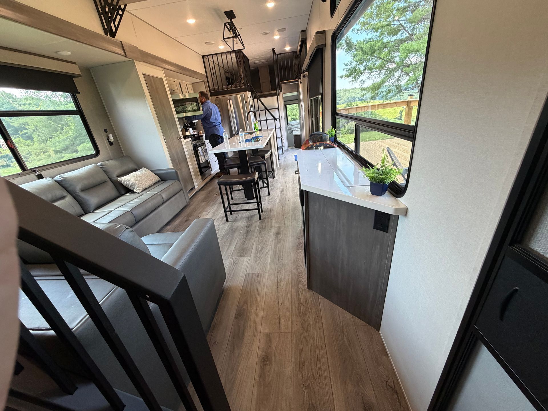 Narrow RV interior with kitchen, sofa, dining table, and large windows overlooking trees