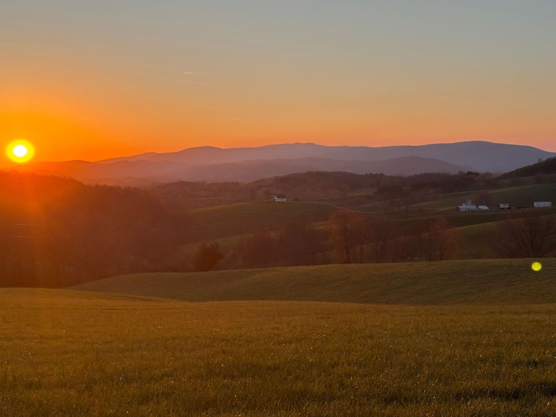 Sunrise over rolling hills and a grassy field, with warm orange sky and distant mountains