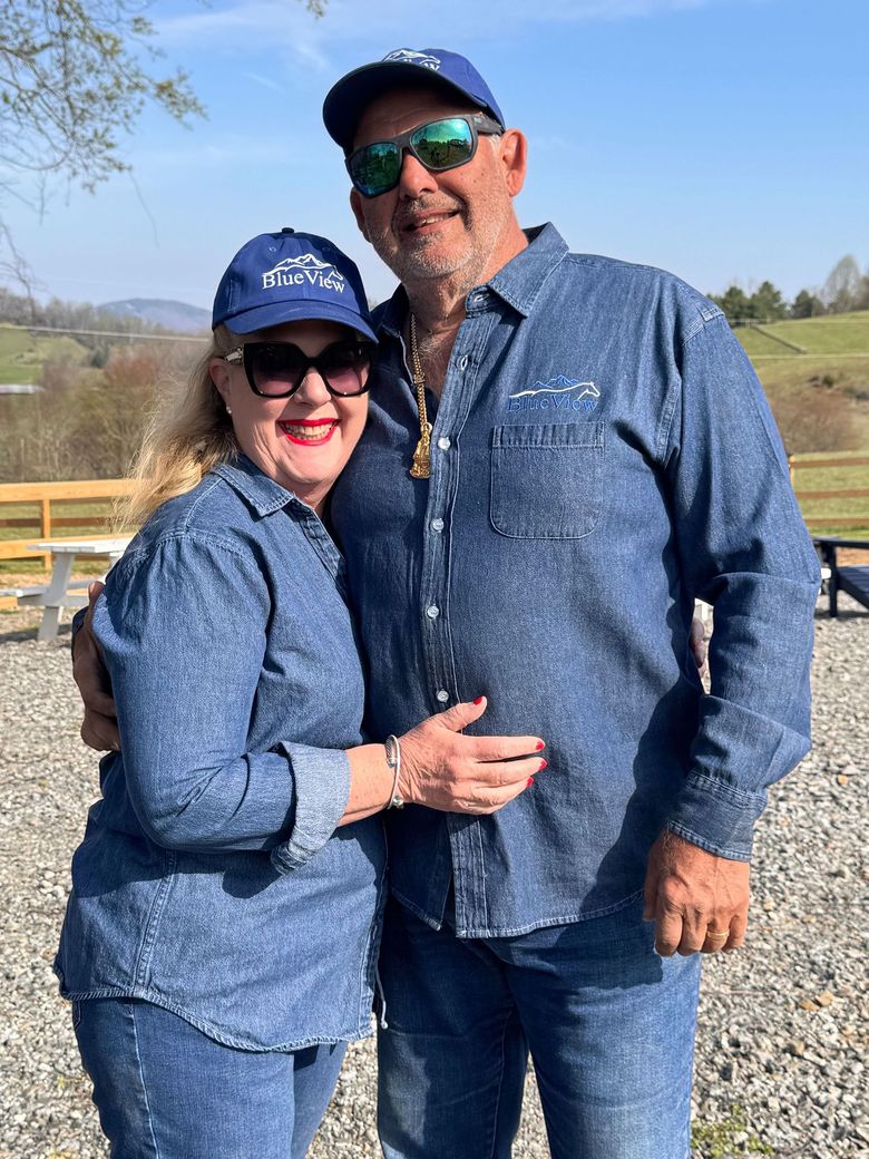 Two smiling people in blue denim outfits and caps standing outdoors by a wooden fence on a sunny day