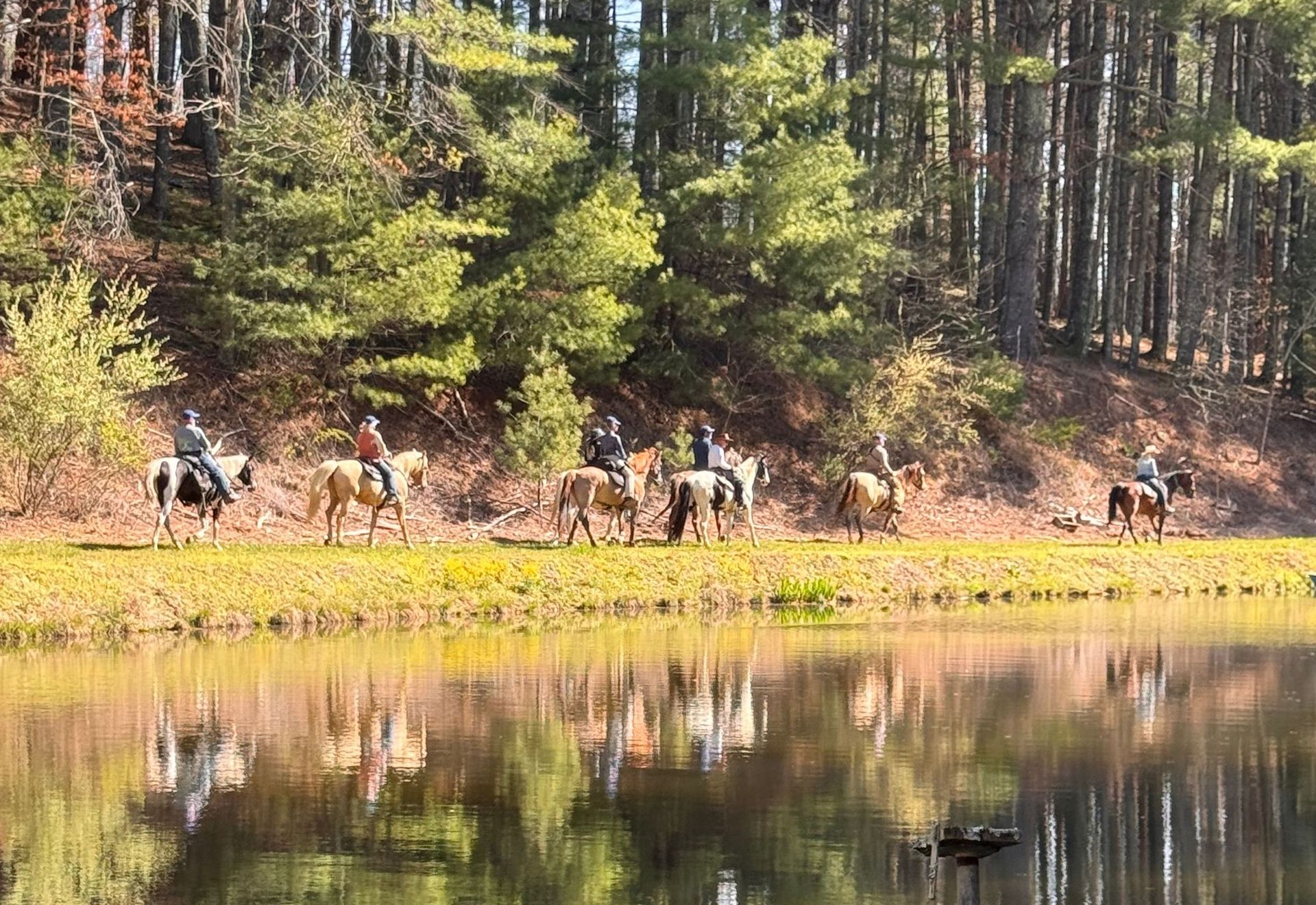 People riding horses along a wooded lake shore, reflected in calm water.