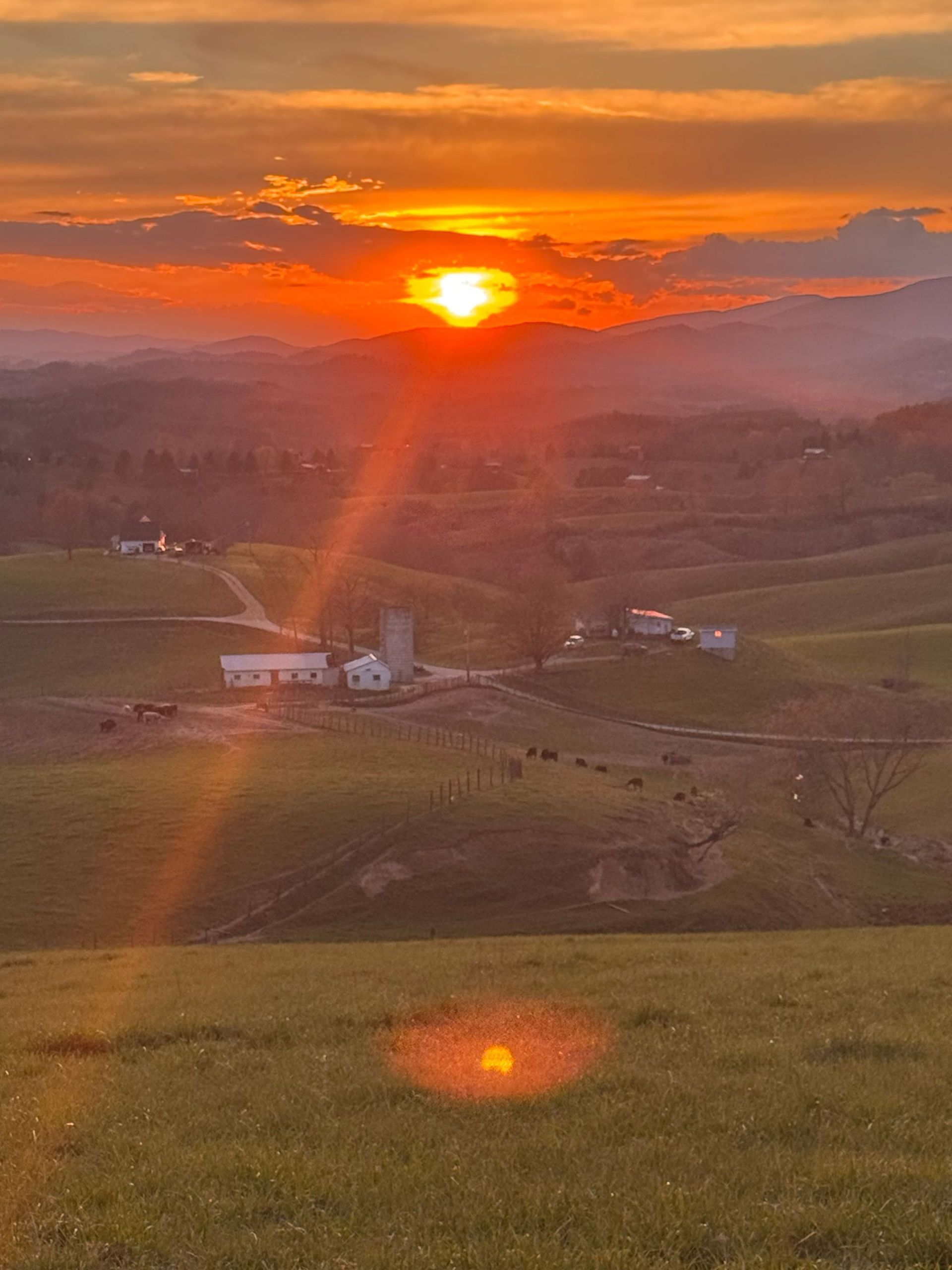 Sunset over rolling green hills, with orange sky and lens flare above a small rural farm.