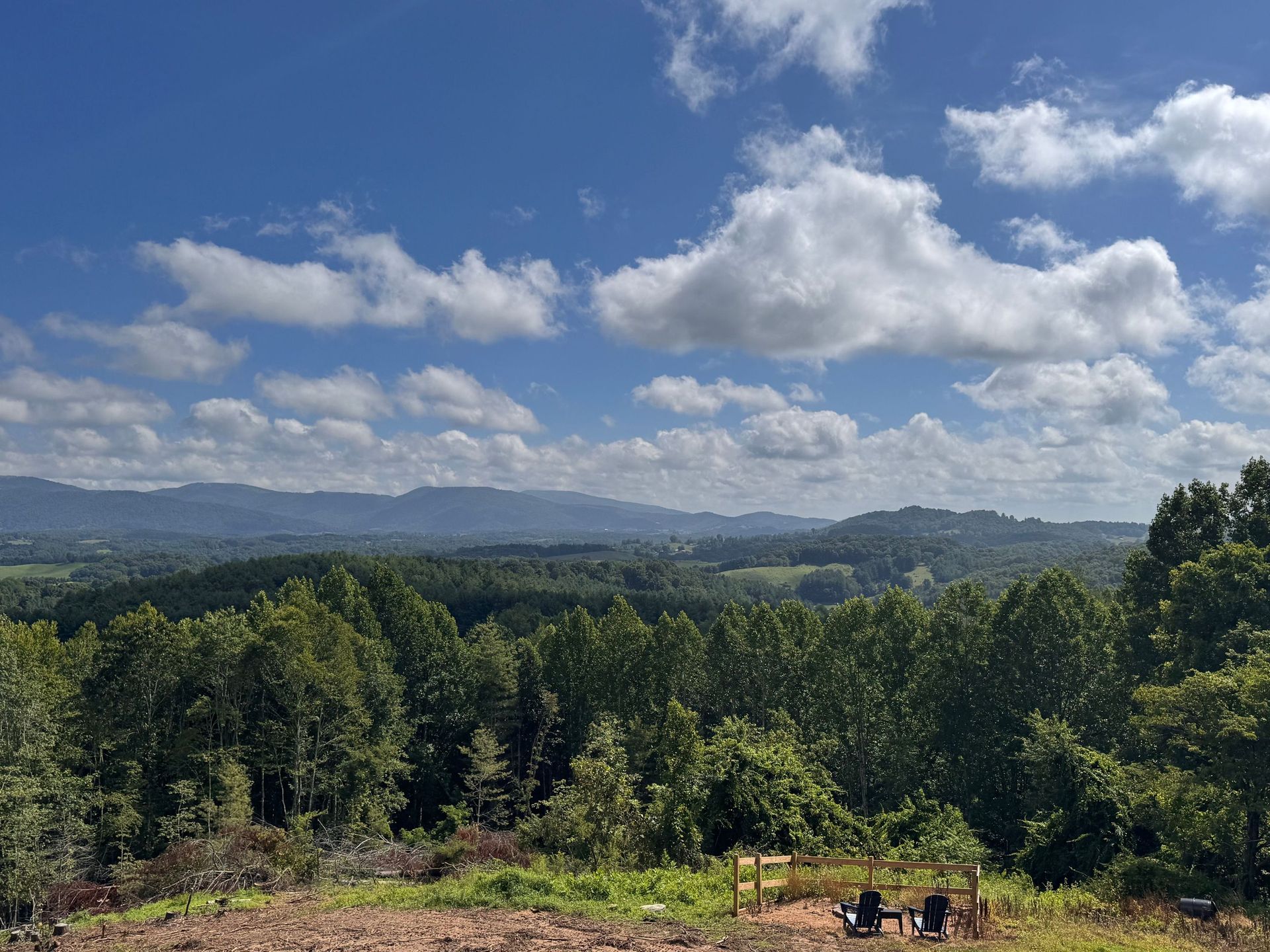 Rolling green hills under a bright blue sky with scattered clouds, viewed over a wooded landscape