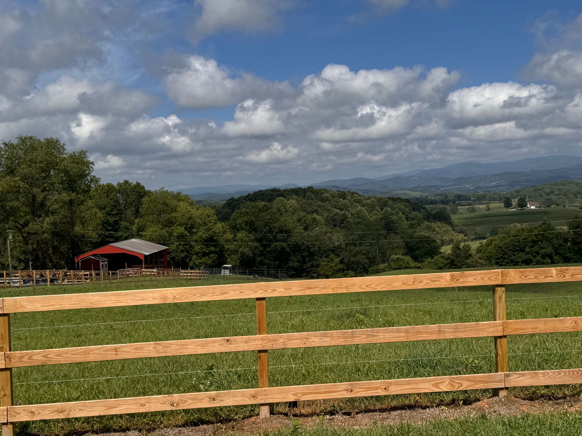 Wooden fence overlooking a grassy field, trees, a red-roofed shed, and distant mountains under a cloudy sky