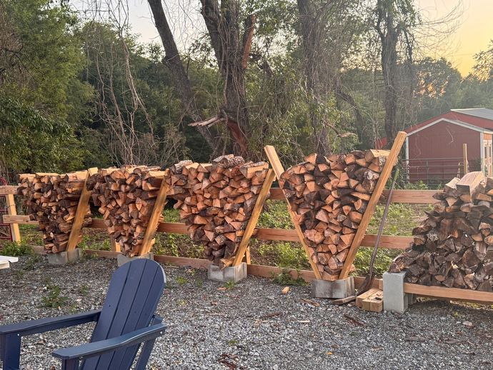 Stacked firewood in wooden racks outdoors, with a blue chair in front and trees and a red shed in the background.