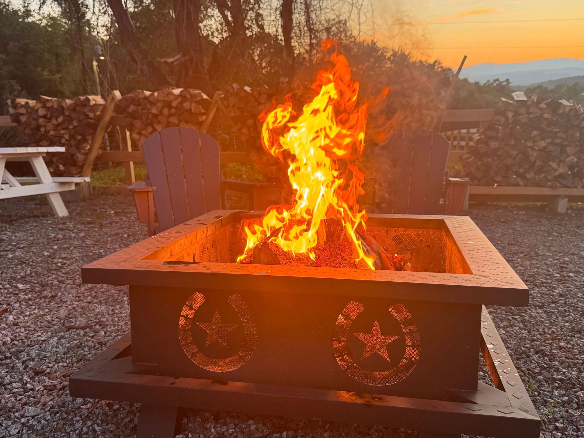 Fire burning in a square outdoor fire pit at sunset, with stacked wood and chairs nearby.