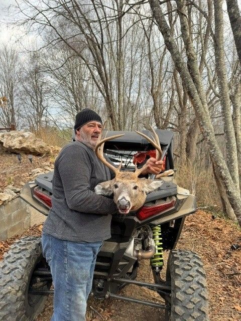 Man standing beside an ATV with deer antlers in a wooded outdoor area