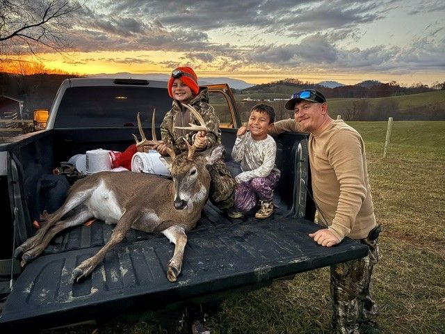 Hunter and two children with a deer in a pickup truck bed at sunset, in a rural field.