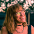 Smiling woman with glasses and long hair, wearing a black top and necklace, against a softly blurred pink background