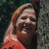 Smiling person with reddish hair leaning against a tree in dappled sunlight