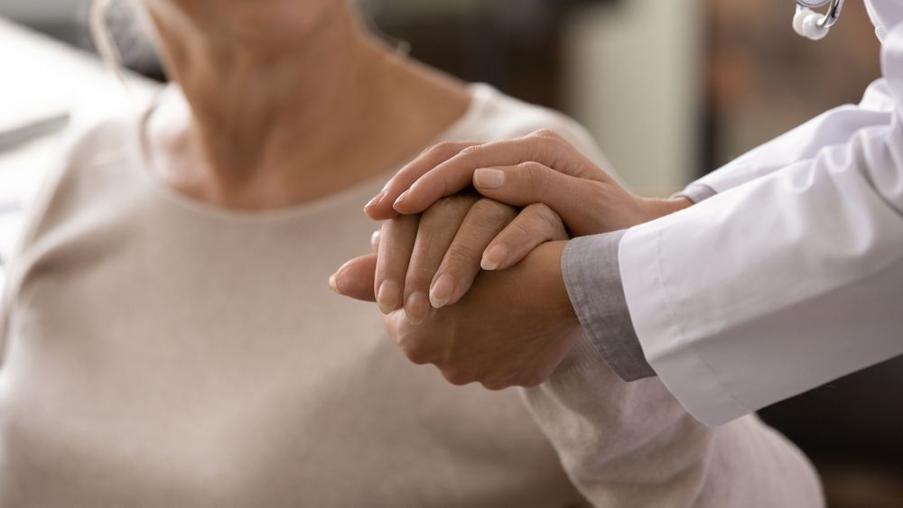 Doctor's hand on a patient's hand for comfort, both hands clasped. Doctor in white coat, neutral tones.