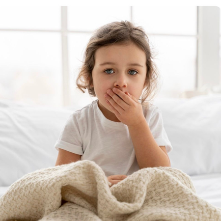Child sitting in bed, hand over mouth, under a beige blanket, in a bright room with white window light