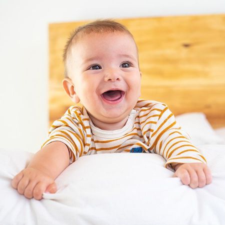 Smiling baby in a striped shirt sitting on a white bed with a wooden headboard background
