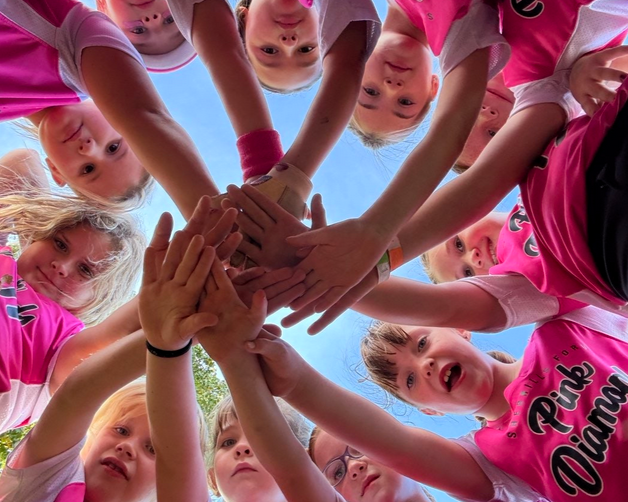 Hands stacked in a team huddle, with children in bright pink shirts leaning in from a circle.