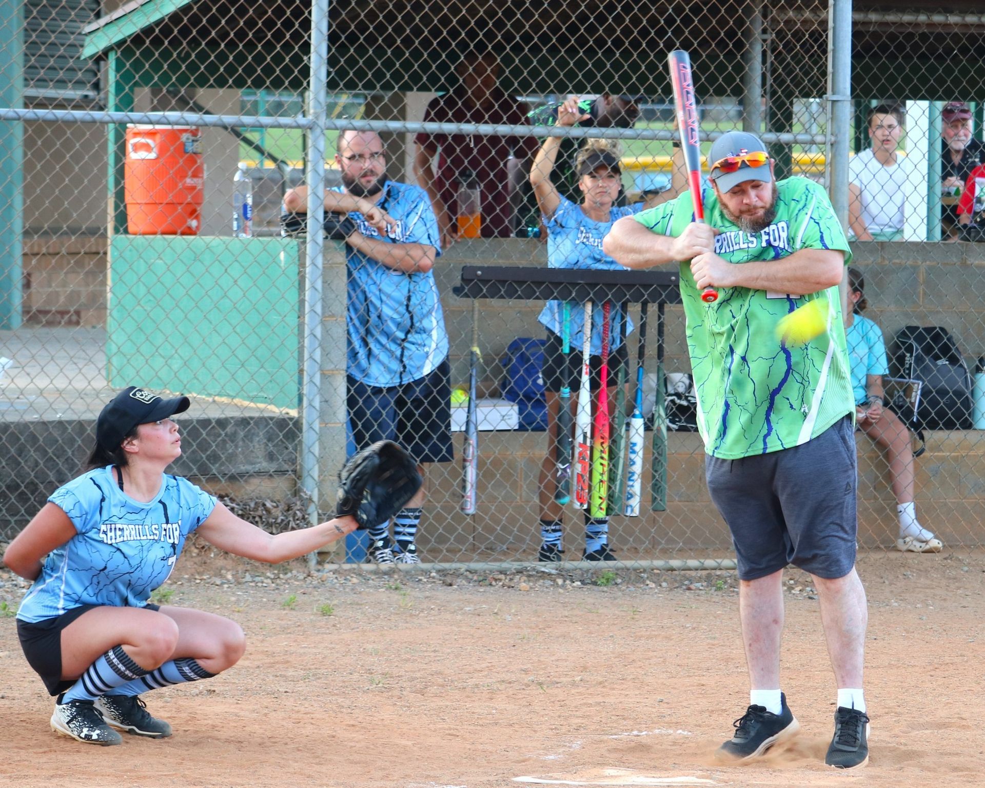 Softball game: batter in green shirt facing catcher. Spectators behind chain-link fence.