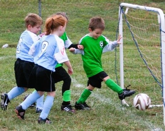 Children playing soccer near a goal, with a goalkeeper in green reaching for the ball.