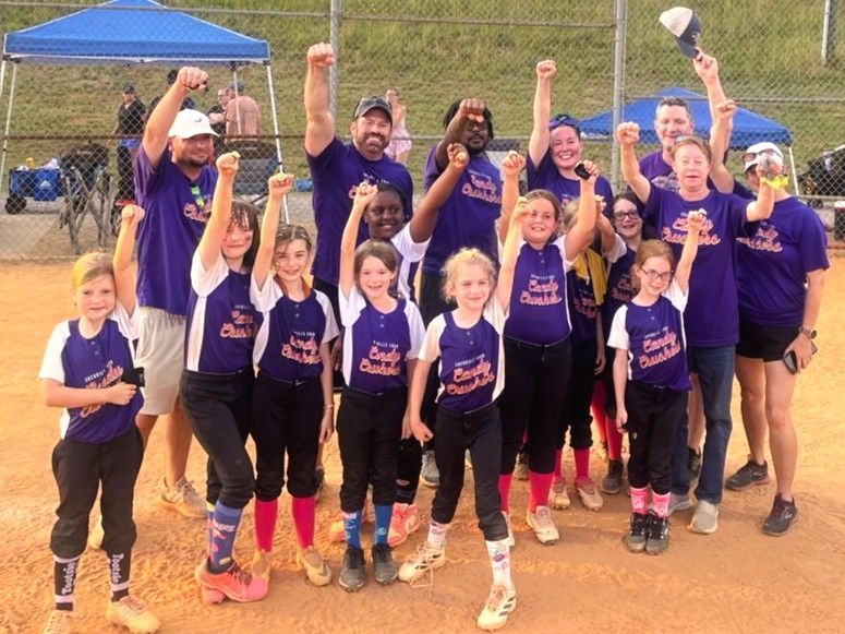 Group of runners in purple shirts posing with raised fists at an outdoor event on a dirt field
