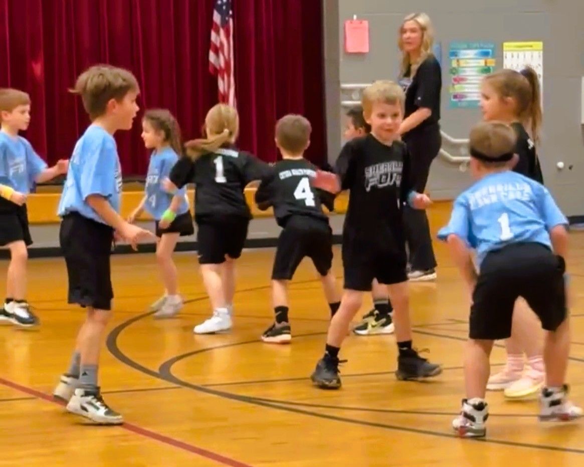 Children playing a sport in a gym. Some wearing black and others blue shirts. A woman watches in the background.