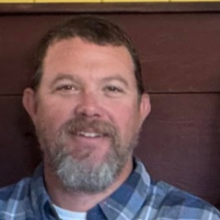 Man with beard, blue plaid shirt, smiling in front of a wood background.