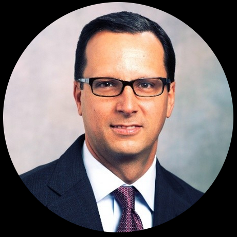 Man in a dark suit and glasses, smiling, set against a light background.