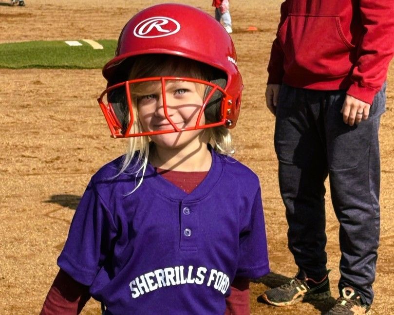 Child wearing a red helmet and purple baseball jersey, smiling on a field.
