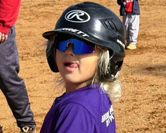 Child in purple shirt and black helmet with reflective sunglasses at a softball field