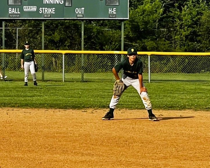 Baseball players on a field. One in ready position, other in the background. Green and yellow uniforms. Scoreboard visible.