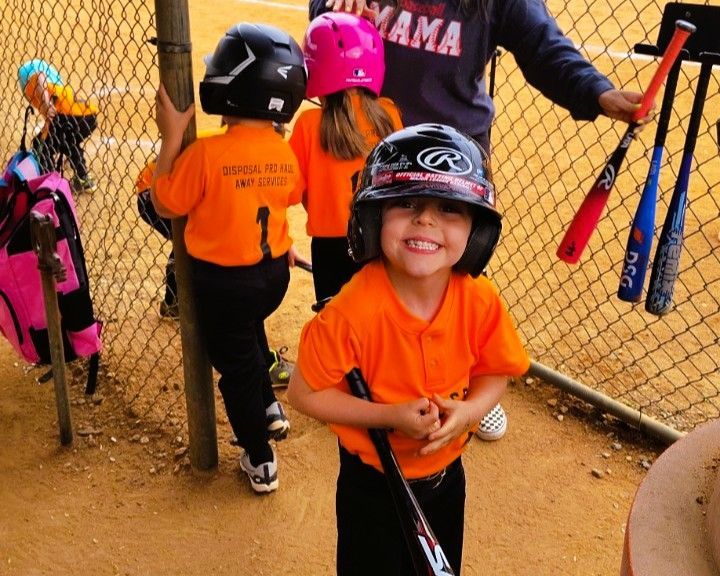 Young softball player in orange uniform smiles, holding a bat, with teammates in the dugout.