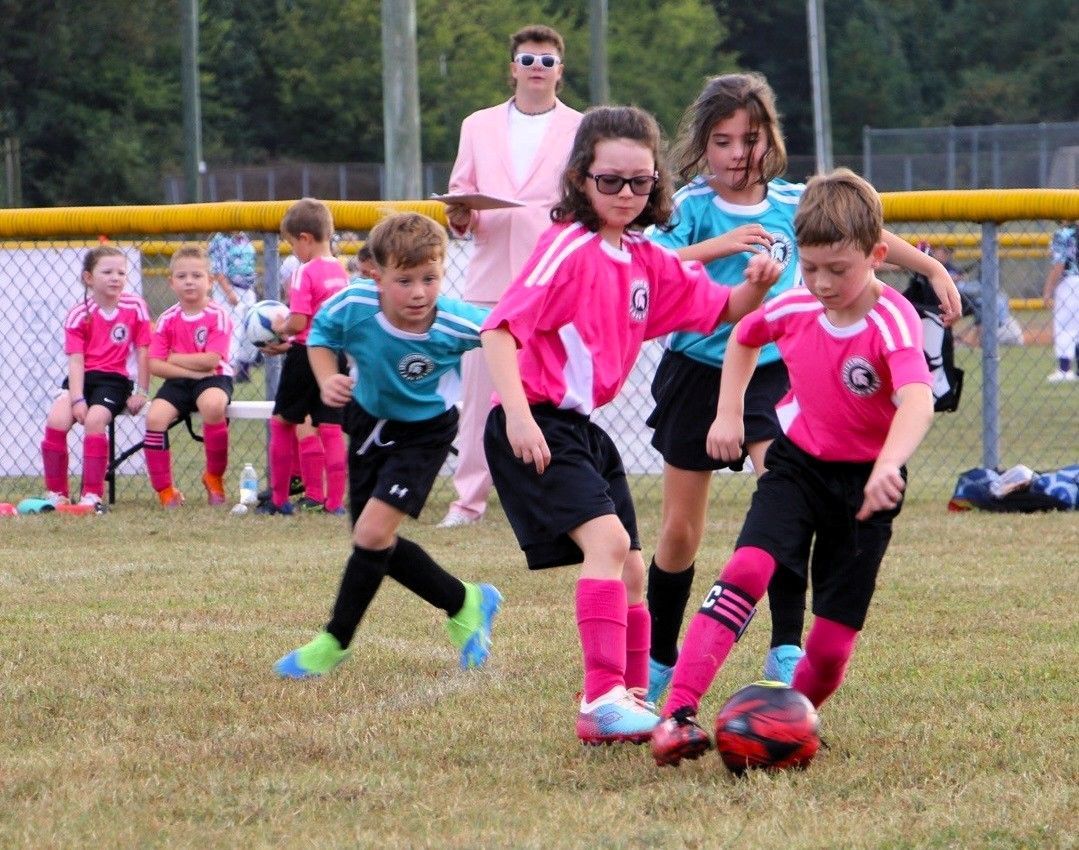 Kids in pink and teal soccer uniforms playing soccer on a field. Coach in pink suit watches.