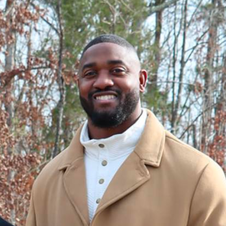 Man wearing tan coat and white turtleneck smiling outdoors.