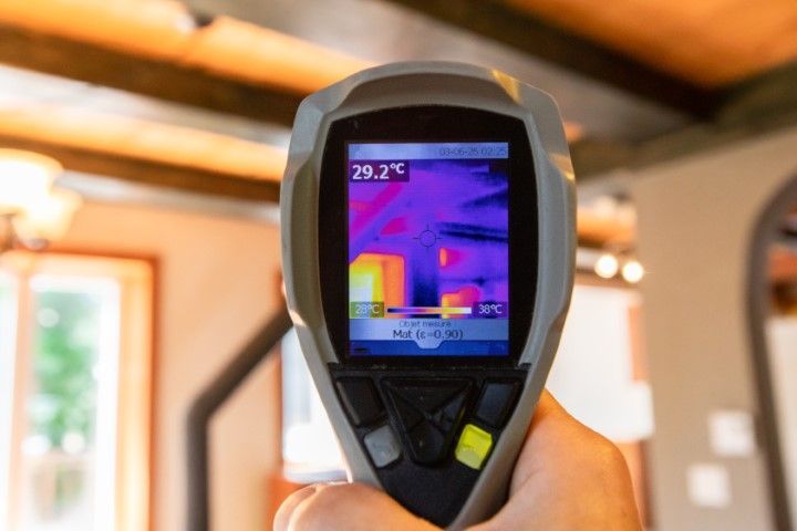 Hand holding thermal camera displaying a heat map of a room, indicating temperature variations.