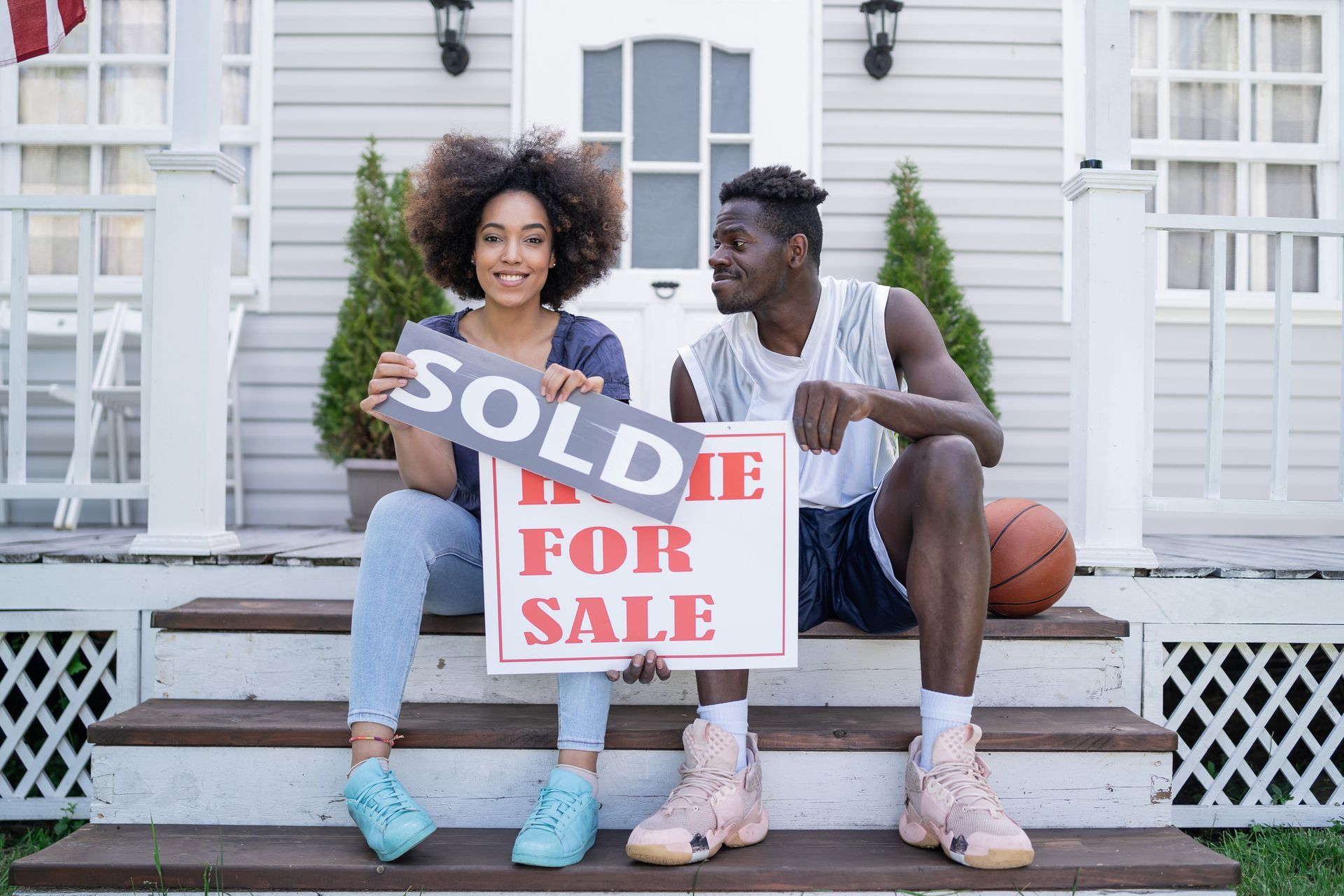 A man and a woman are sitting on the steps of a house holding a sold sign.