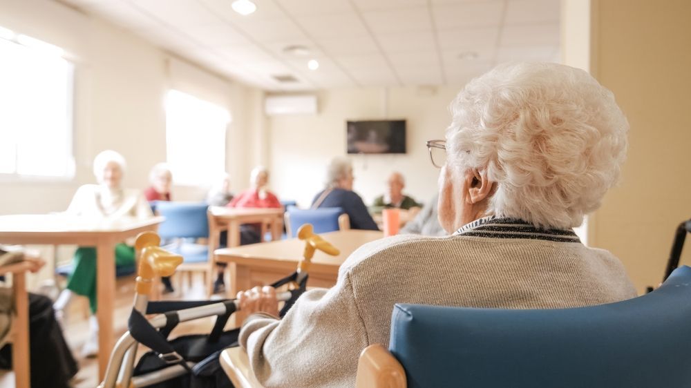 Elderly woman in a wheelchair facing a room of other elderly people; in a senior living facility.