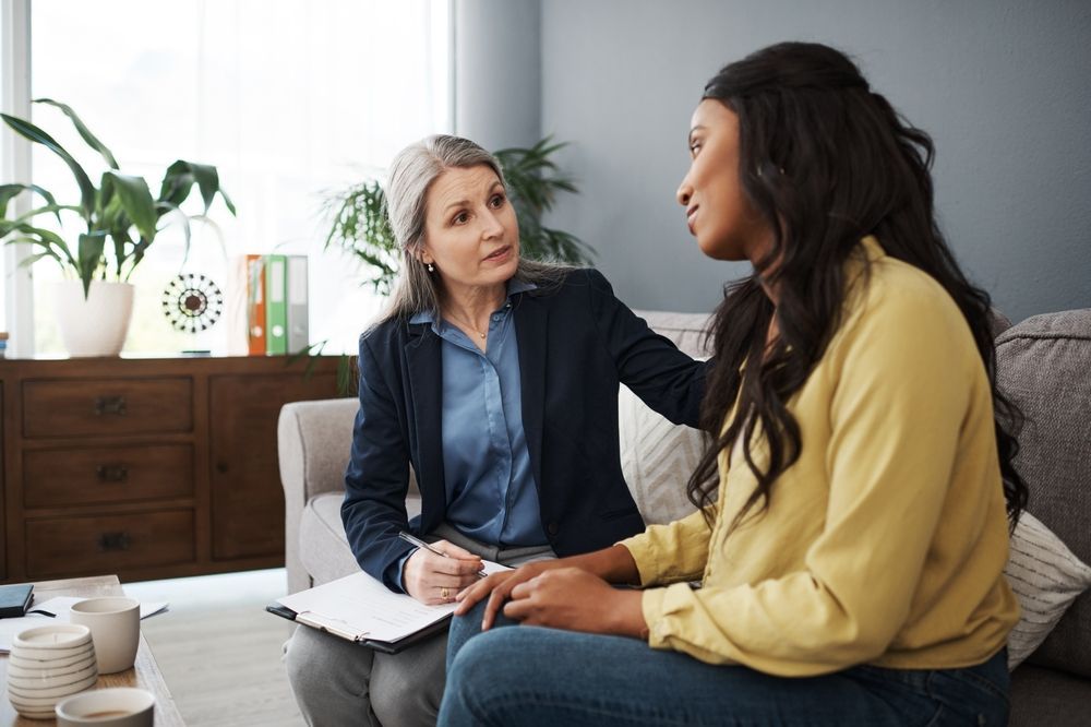 Woman comforts a woman in a counseling session. They are in a living room, and the counselor is taking notes.