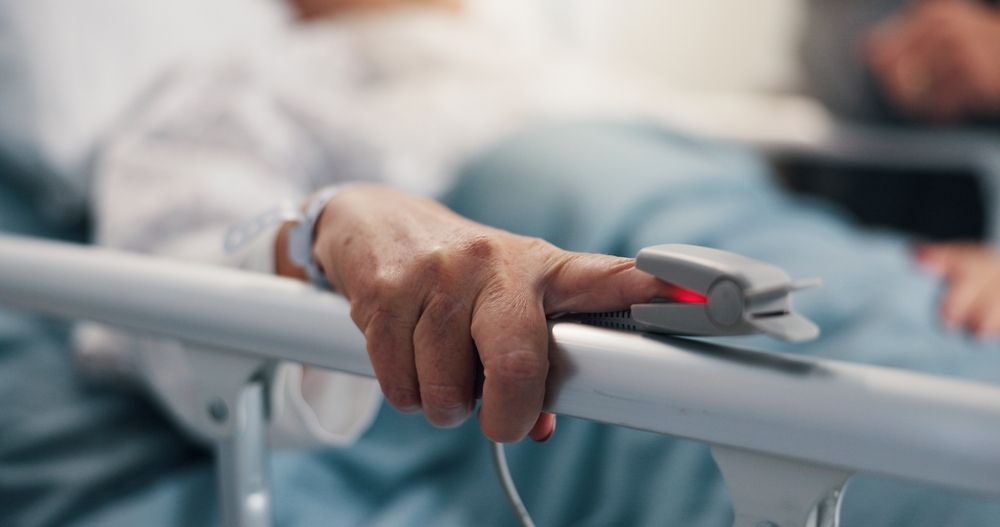 Person's hand with a pulse oximeter clipped to a finger, resting on a hospital bed rail.