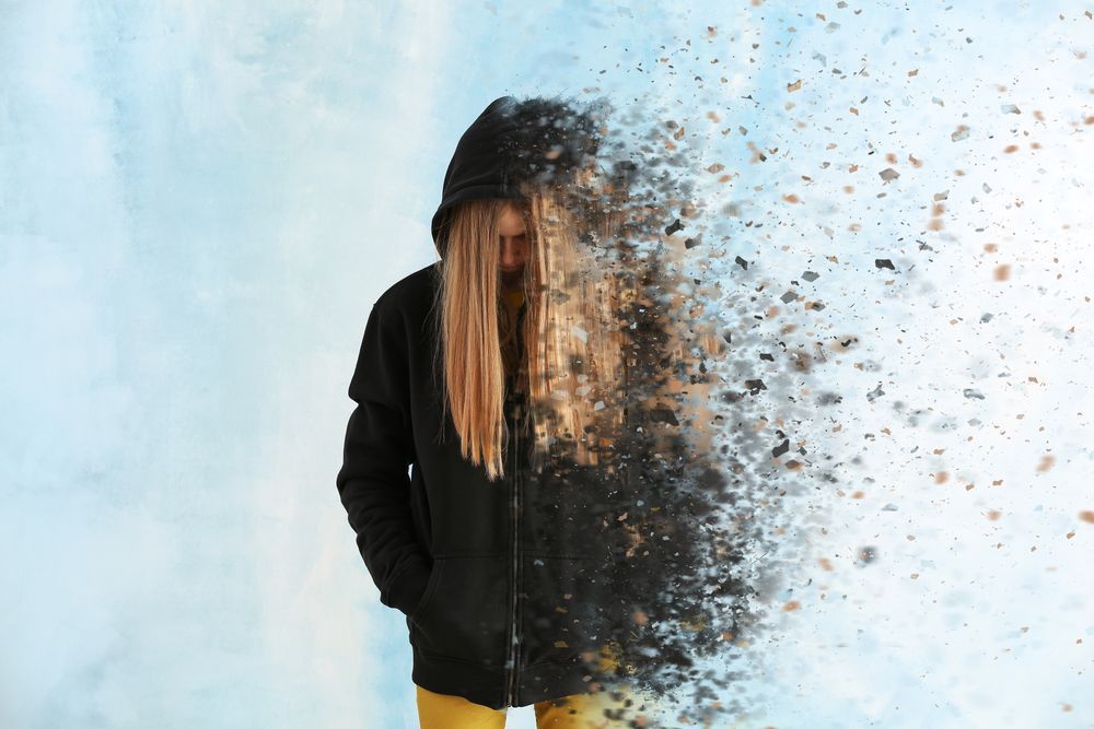 A person in a black hoodie and yellow pants is dissolving into particles against a light blue background.