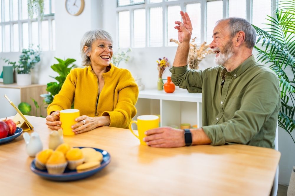 Smiling couple at a table, woman in yellow, man gesturing. Kitchen setting, mugs, breakfast food.