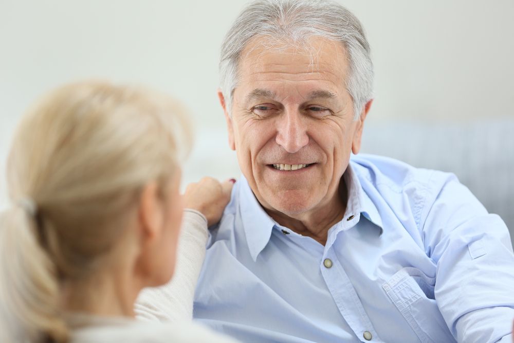 Smiling older man in a blue shirt with a woman; arm on his shoulder. Indoors, casual setting.