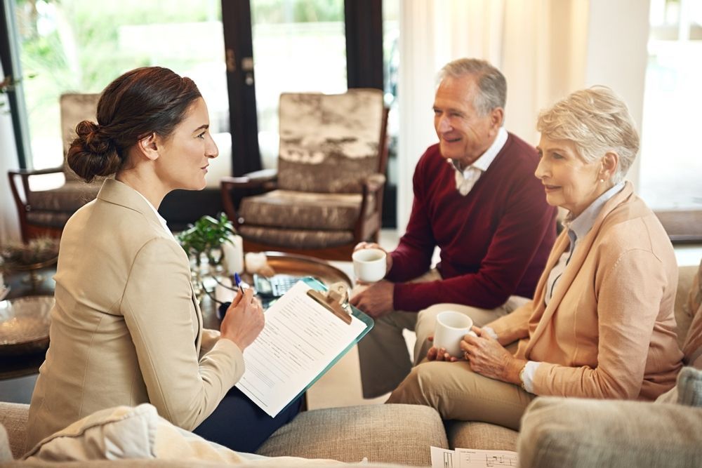 Woman in blazer reviews paperwork with smiling senior couple on a couch.